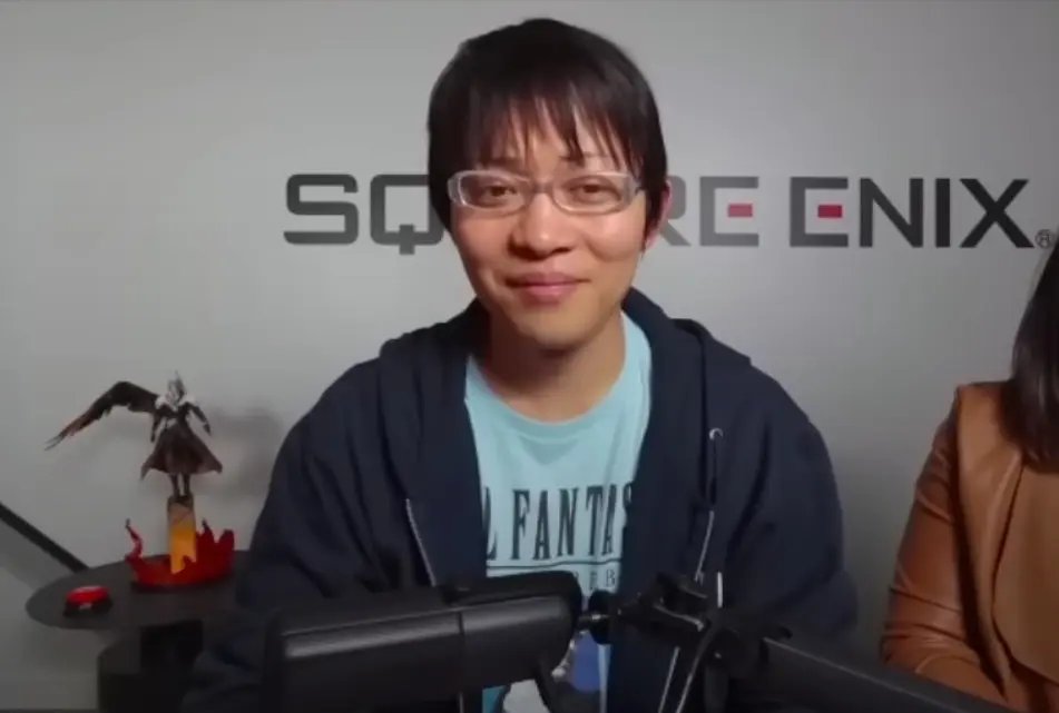 Naoki Hamaguchi v brýlích a modrém tričku sedí před stěnou s logem Square Enix.