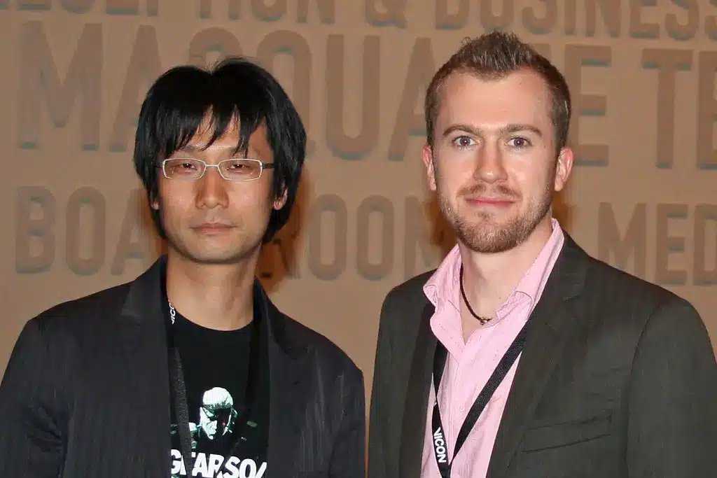 Hideo Kojima v brýlích a černém saku stojí vedle jiného muže.